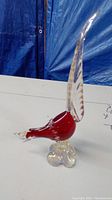 Front and side views of an art glass pheasant sculpture with a red body and a tall clear glass tail on a clear base. The sculpture appears damaged.
