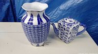 Blue and white ceramic vase with a flared shaped rim and grid pattern beside a teapot-shaped ceramic votive holder with lid and handle, both placed on a table against a blue backdrop.