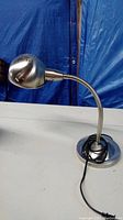 Metal desk lamp standing on table with flexible neck extended and power cord coiled at the base.