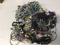 Top view of large tangled pile of costume jewelry including multi-colored beads, faux pearls, metal chains, and black coiled accessory.