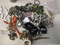 Photo showing a large pile of assorted costume necklaces and bracelets, including faux pearl white and black beads, orange and purple beads, various gold-tone chains, and other mixed bead styles, all tangled together on a flat surface.