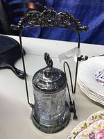 Front view of silver plate and cut glass pickle jar with ornate metal handle and lid, and attached tongs.