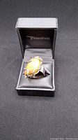Ring displayed in a black jewelry box with 'slamad goldsmithing' branding, featuring a large honey-colored amber stone in a silver setting.