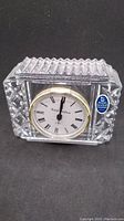 Front and angled view of the rectangular crystal clock showing textured sides and top with clear crystal casing and round clock face with Roman numerals and gold-tone bezel.