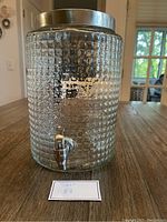 Front view of the textured glass beverage dispenser showing metal lid and spigot on wooden table.