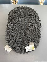 Set of eight black round woven placemats stacked together on a table with visible lot tag.