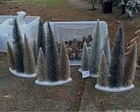 Five bottlebrush tabletop Christmas trees arranged on white bases with natural pinecones in a container behind them, all displayed outdoors on ground covered with pine needles.