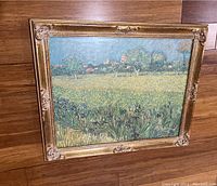Front view of a rectangular gold ornate frame with a landscape painting showing a field, iris flowers, trees, and buildings in the distance.