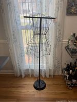 Full view of a 53-inch tall black metal wireframe dress mannequin in front of a sheer curtain background with wood floor and nearby liquor bottles.