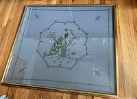 Full view of framed hand-embroidered peacock artwork on cloth with gold frame lying on hardwood floor.