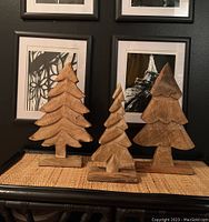 Three wooden stylized tree decor pieces displayed together on a surface with black wall and framed pictures in the background.
