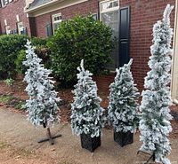 Four pre-lit flocked artificial Christmas trees outdoors against brick wall and shrubbery, showing overall size and condition.