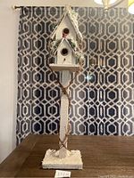 Full view of the decorative bird house standing on a table with a patterned curtain background, showing the entire structure including base, post, and top houses.