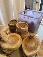 Wood slab slices, wooden bowls, and card holders seen stacked beside a plastic basket on a table with a cream curtain background.