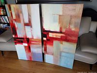 Two abstract canvas prints leaning against a couch on a wooden floor. Both feature similar geometric abstract designs in red, white, beige, and blue hues.