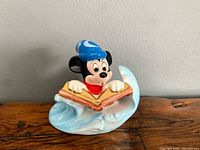 Front view of Mickey Mouse figurine wearing sorcerer's blue hat and red robe, holding an open book, resting on a blue and white wave base.