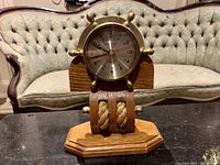 Nautical clock on wooden and rope stand shown on table with sofa background.