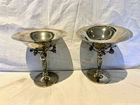 Two silver-plated decorative dishes on stands, showing front and side angles with grapevine embellishments on stem.