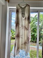 Full length photo of white kaftan with brown leaf-like patterns draped on hanger in front of window with garden view