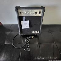 Front view of black Eleca guitar amplifier on black surface showing control panel knobs and input, power cord coiled in front.