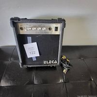 Front angle view of black Eleca EG-10J guitar amplifier with control knobs and power cord on a black surface.
