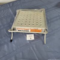 Little Giant work platform accessory placed on blue fabric surface, showing perforated metal platform and metal frame with capped legs. Front view.
