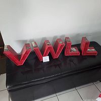 Photo showing four large red metal sign letters V, W, L, L placed on a black bench. Letter tops have metal mounts. Visible wear and aging on paint.