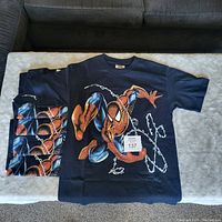 T-shirts displayed folded and one unfolded showing the Spider-Man graphic design with webbing and action pose on dark blue.