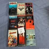 Top-down view of 9 VHS horror movie tape cases arranged in a 3x3 grid on a fabric surface.