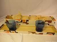 Six blue coloured vintage Boussu drinking glasses arranged in two rows inside a retro patterned box with tissue paper wrapping some glasses.