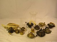 Overall lot view showing multiple brass and metal oil lamp burners, two glass chimneys, and fabric wicks
