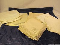 Lot of assorted vintage linens laid out on a dark cloth background, showing different folded pieces with crocheted and plain edges.
