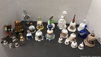 Photo showing collection of approximately 25 bells arranged in rows on a tabletop, varied sizes from 6 inches to about 1 inch. Bells made of porcelain, glass, and metal with painted and metallic designs. Some have animal or figurine handles.