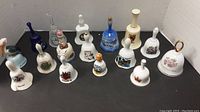 Front and overview of approximately 15 porcelain and glass decorative collectible bells arranged on flat surface