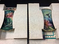 Two Bradford hummingbird decorative plates partially visible inside original white cardboard boxes showing floral and bird designs with green and blue backgrounds.