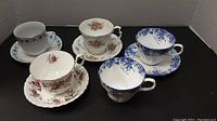 Front view of the five cups and saucers on black surface showing floral patterns and types of cups and saucers.