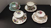 Four porcelain cups and saucers arranged with floral patterns and gold trim on a dark surface.