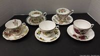 Five vintage floral porcelain cups and saucers grouped on black background with white cardboard sides.