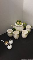 Daisy tea pot with lid, four mugs, and white ceramic salt, pepper, and spice jars on black surface.