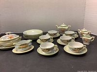 Full view of all tea party dish pieces for dolls featuring teapot, cups, saucers, plates, covered serving dish, creamer, and sugar bowl arranged on dark surface with white backdrop.
