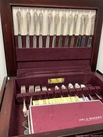 Open flatware cabinet showing burgundy velvet interior lining with 12 place settings of silverplate knives, forks, spoons, and serving pieces in Enchantment pattern by 1881 Rogers Oneida.