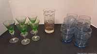 Full set of 5 green stemmed sherry glasses, 6 blue textured tumblers, and 1 tall gold decorated glass shown together on black surface with white background.