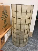 Front view of cylindrical shell lamp shade with metal grid frame placed on carpet next to boxes.