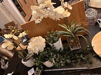 Seven different artificial plants and flowers displayed on a table including white orchids, a white peony flower, green trailing vine plant, variegated succulent, and snake plant in clear glass vase.