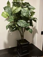 Artificial plant with broad green variegated leaves and multiple stems in a black square planter, viewed from the front.