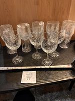 Photo showing set of eight crystal wine glasses lined up on a dark surface against wood background. Visible ruler in front for scale.