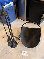 Photo of fireplace tool set including stand, poker, brush, shovel, and metal ash bucket with handle placed on floor.