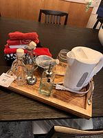 Wooden tray on dark wood table holding white electric kettle, glass containers, glass soap dispenser, and red kitchen linens