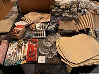 Photo showing a table covered with a wide variety of kitchen items, including jars, napkins, placemats, spoons and silver plated servers and cutlery.