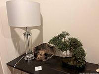 Table lamp, stone decor slab, and artificial bonsai tree on black surface against white wall.